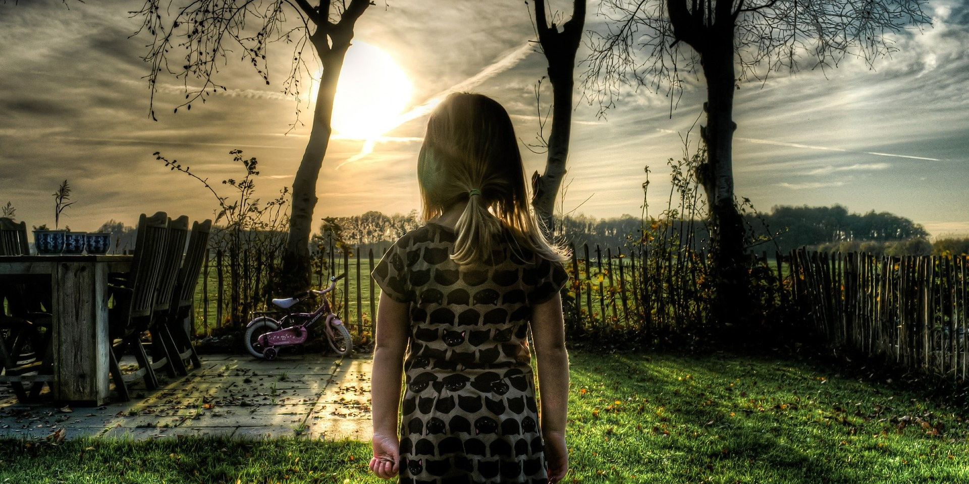 Ein kleines Mädchen steht alleine im Garten und schaut in den bewölkten Himmel 